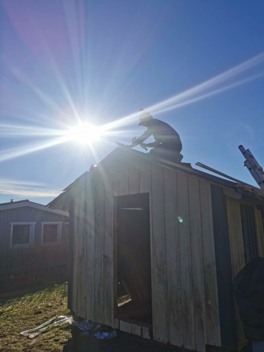 Person arbetar på taket till ett litet uthus under en solig himmel.