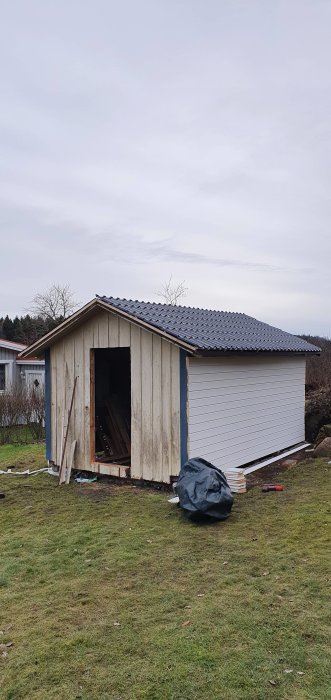 Ett halvfärdigt träuthus med nytt tak och öppen vägg, byggmaterial på marken, under pågående renovering.