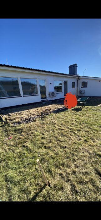 Grävmaskin och skottkärra nära ett hus, förberedelse för altankonstruktion med synliga plintpositioner i sanden.