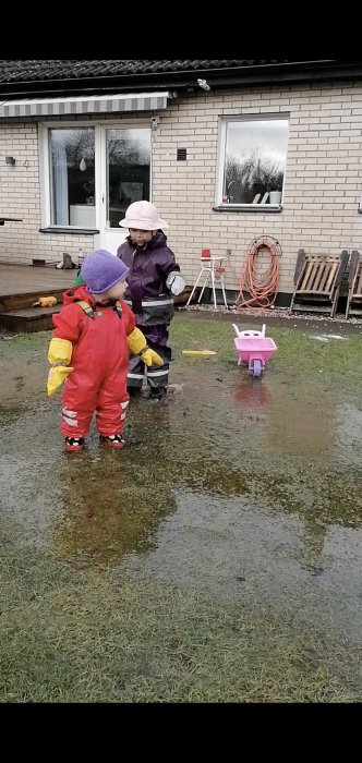 Två barn i regnkläder leker i en vattensamling i trädgården framför ett hus med synlig fönster och trädgårdsslang.