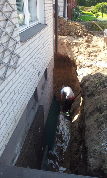 Person utför dräneringsarbete vid husgrund med uppkopad jord och synlig fuktspärr.