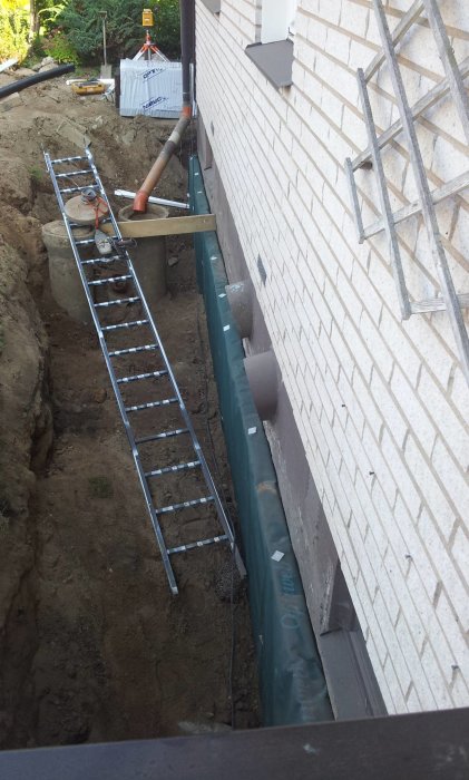 Dräneringsarbete längs husvägg med öppnad schakt och ny dräneringsledning, stegar och byggmaterial synliga.
