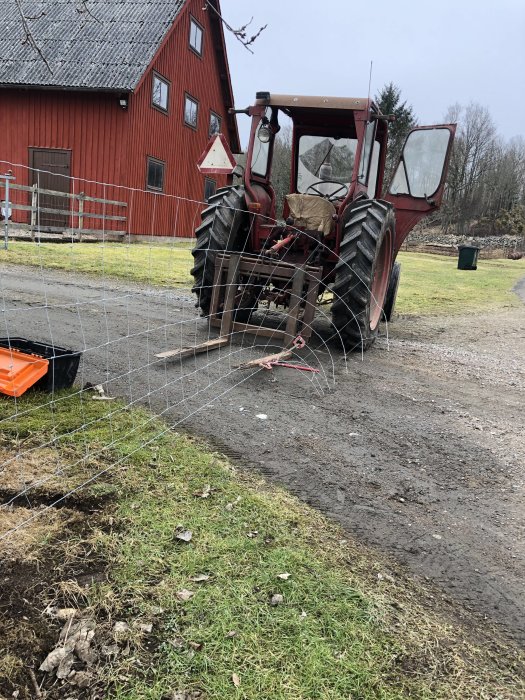 Traktor som håller i ett nystan av staket vid en grusväg med ett rött hus i bakgrunden.