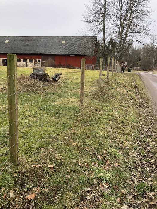 Nyligen uppsatt staket längs en vägkant med en röd lada i bakgrunden.