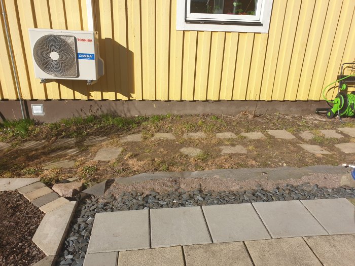 En ojämn gräs- och jordremsa mellan ett gult hus med luftvärmepump och en stenlagd gångväg.