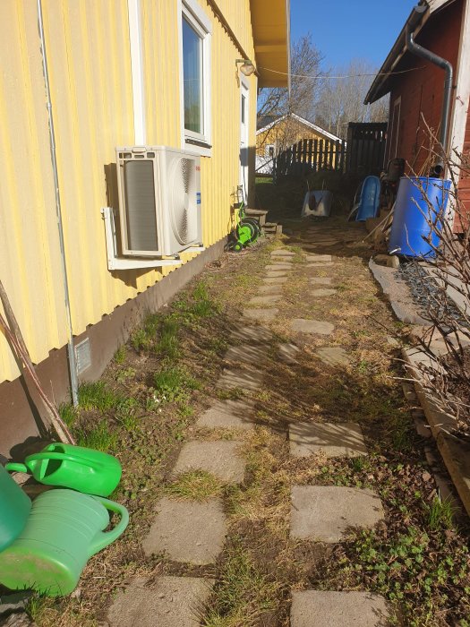 Gångränna längs husvägg med ojämna plattor och gräs, behöver renovering.