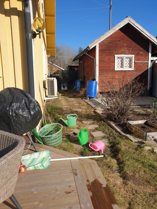 Trädgård med en nybyggd altan som leder till en outgrävd passage med gräsklumpar intill ett rödfärgat förråd.