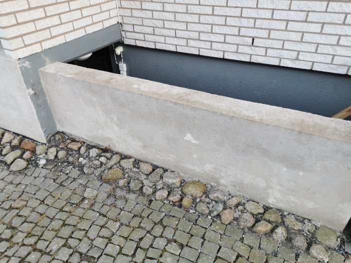 Betongtrappa och mur med synliga skador och släpp i putsen framför tegelhus och stenbelagd mark.