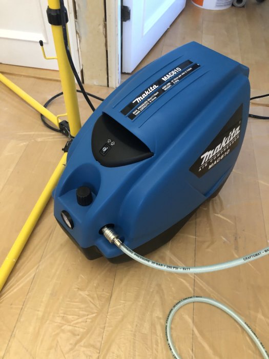 Blå Makita luftkompressor för dyckertpistol uppkopplad med gul luftslang på trägolv.