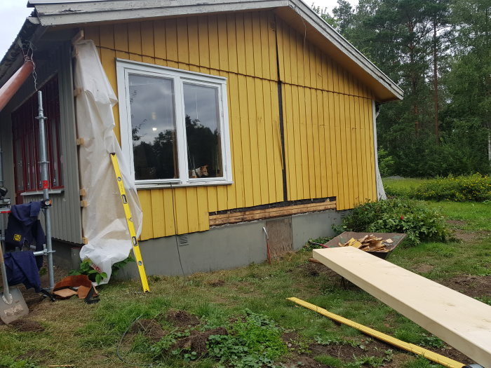 Gult hus med pågående renovering, inklusive skyddstält, stege, borttagna fönster och byggmaterial på marken.