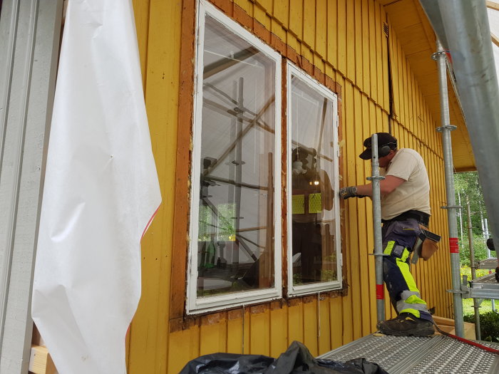 Person i arbetskläder tar bort ett fönster från en gul trävägg på en byggställning.