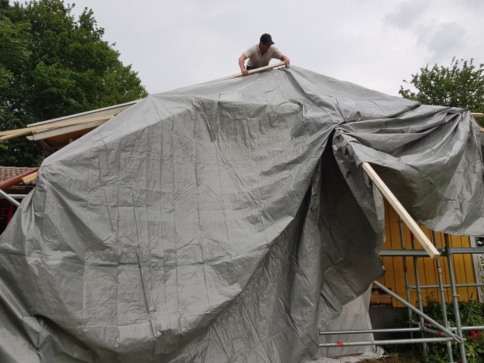 Person på taket justerar grå presenning över en byggställning vid husrenovering.