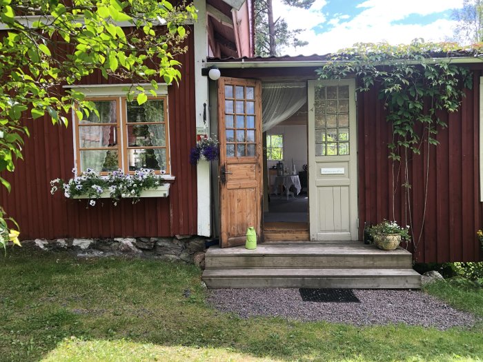 Röd sommarstuga med begagnade pardörrar, utan entrétak, omgiven av grönska och en blomsterfönsterkarm.