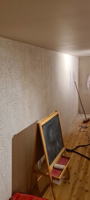 Renoveringsrum med kritmoln på svart tavla, ojämn vitmålad vägg och trägolv med renoveringsmaterial.