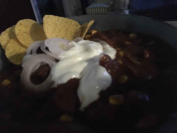 Skål med chili, grekisk yoghurt, picklad lök och nachos, bilden är mörk och suddig.
