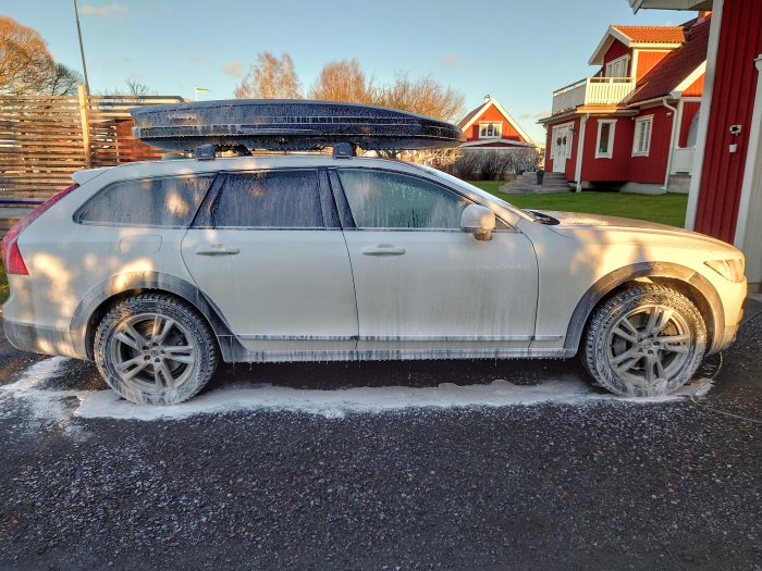 Halvt snötäckt kombibil med takbox parkerad på en solbelyst gata.