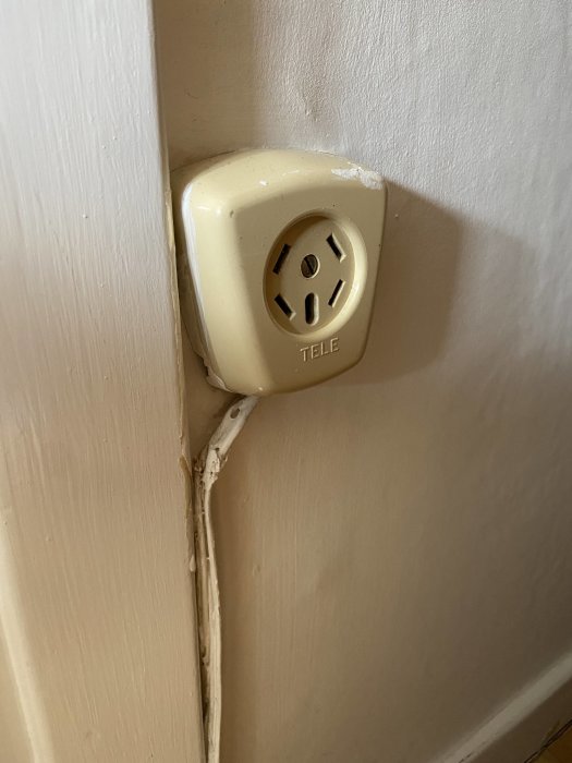 Gammalt beige telefonuttag märkt "TELE" på en vit vägg med frilagd gammal kabel.