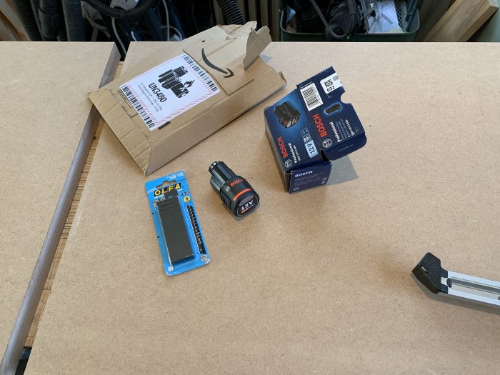 Nytt 3Ah Bosch-batteri, kartong och fraktpaket på verkstadsbord, med kniv och maskindel i förgrunden.