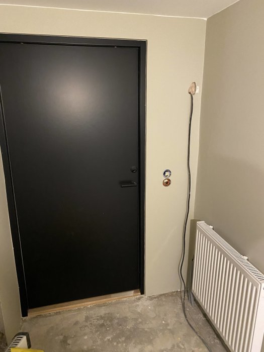 Renoverad källarhörna med ny svart dörr, nymålad vägg och betonggolv med en temporär elkabel och radiator.