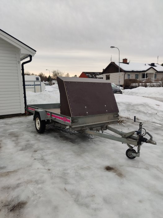 Nyfärdigställd släpvagn med mörk kåpa parkerad på en isig uppfart med bostadshus i bakgrunden.