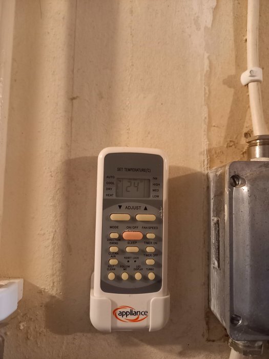 Väggmonterad fjärrkontroll för värmepump med display som visar 24 grader, olika knappar och appliance-logotyp.