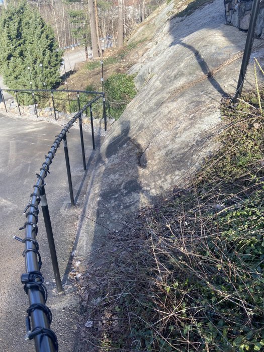 Uppfart intill ett stort berg med en svart metallstaket, torra buskar och träd i bakgrunden.