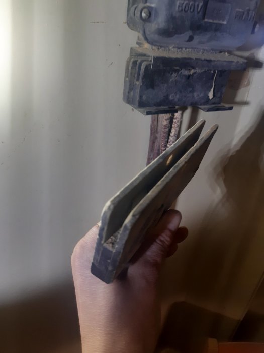 Hand håller ett användt och slitet verktyg framför en gammal säkerhetsbrytare på en vägg.