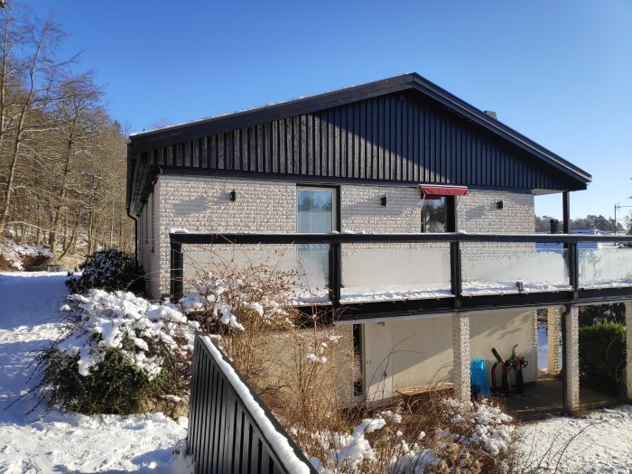 70-talshus med vit tegelfasad och svart panel, altandörr med vita karmar och svart foder, snötäckt mark.
