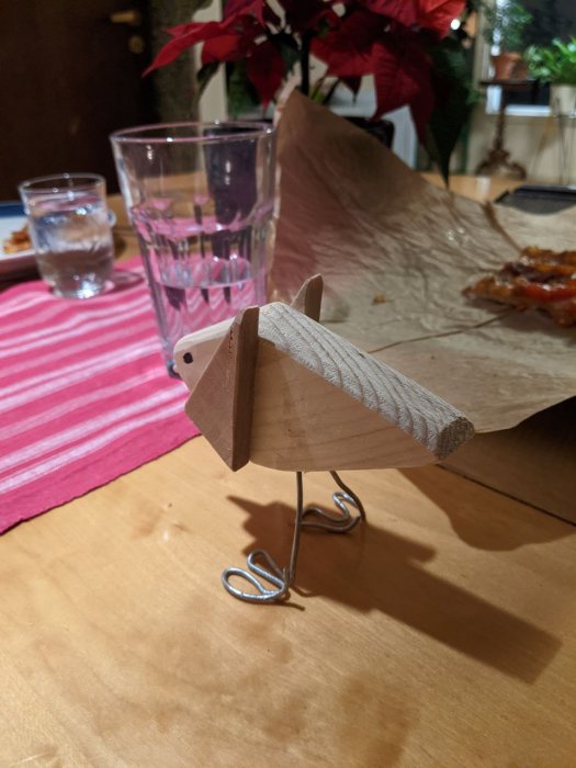 Hantverk av en träfågel med metallben på ett bord, framför växter och glas.