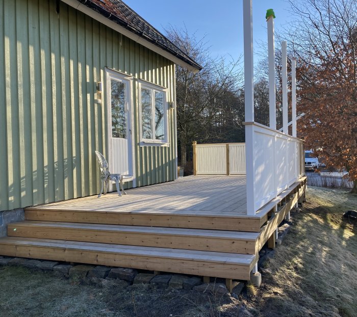 En träaltan i anslutning till ett grönt hus med vita detaljer, med en vit stol och plats för ett framtida altantak.