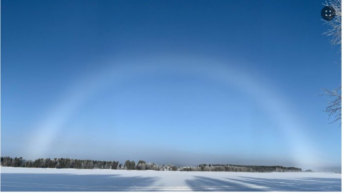 Vit regnbåge ovanför Ångermanälven med snötäckt landskap och klarblå himmel.