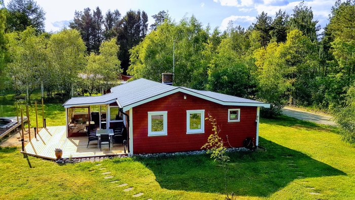 Rött hus med nytt tak och trädäck, omgivet av grönska under solig dag.