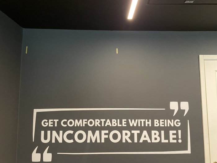 Vägg med text "GET COMFORTABLE WITH BEING UNCOMFORTABLE!" och två gula tejpremsor som markerar reglar.