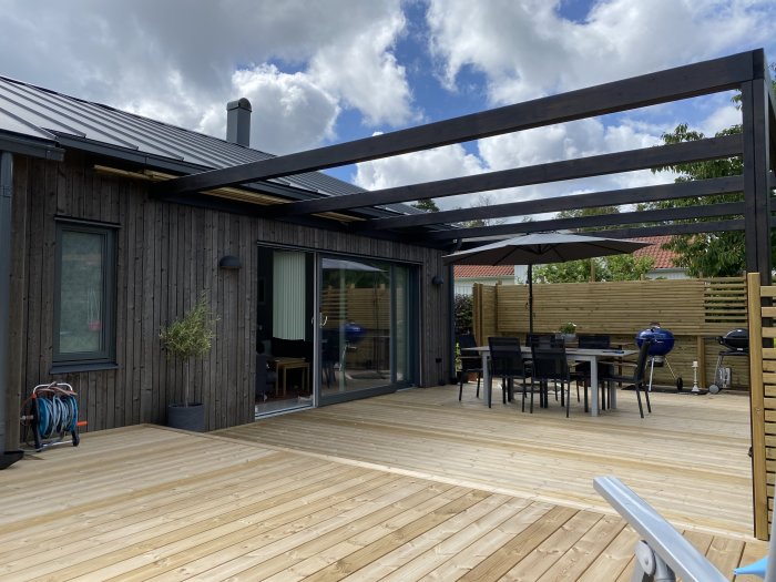 Nybyggd träterrass med möbler och pergola vid ett modernt hus med glasdörrar och träfasad.