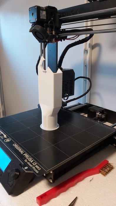 3D-skrivare som skriver ut en vit plastkomponent med överhängande läpp, med verktyg bredvid.