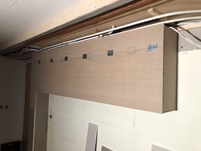 Uppstärkt brun MDF-panel med sex infällda gångjärn och utrymme för drivdon, installerad under tak med synliga kablar.