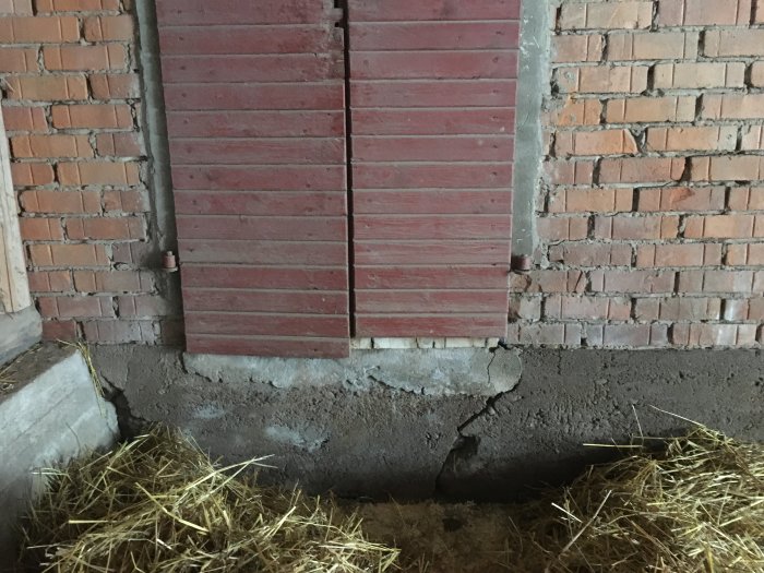 Skadad tegelvägg och träpanel i en lada med sprickor och skador nära betonggolv och hö.