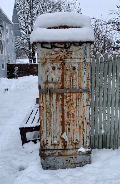 Rostig gammal elskåp täckt med snö framför ett staket, i en trädgård under vintern.