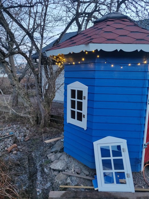 Ett blått hus i Mumin-stil med rött tak och ljusslinga under takfoten, fönster står lutat mot väggen.