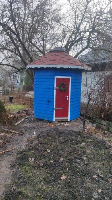 Blått rundat Muminhus med röd dörr och dörrkrans, röd takbeklädnad mot en trädgårdsbakgrund.