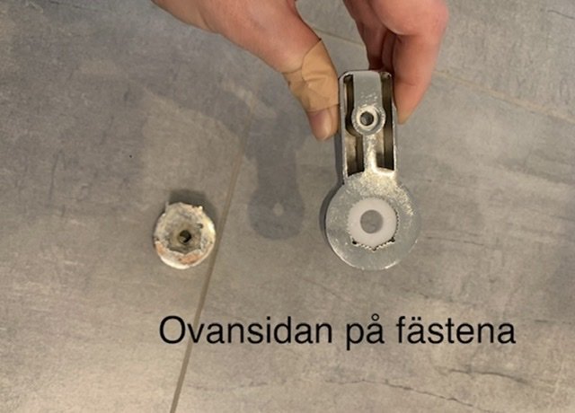 Hand håller en trasig duschdörrsfäste med notering "Ovansidan på fästena".