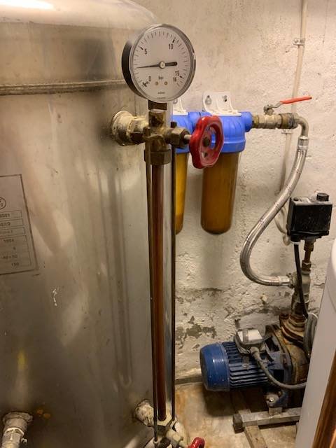 Vattenpump och hydrofor i pannrum med rörledningar, manometer och filtreringssystem.