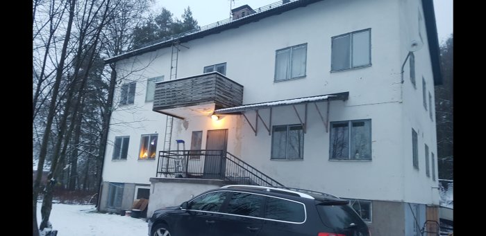 Tvåvåningshus med snötäckt balkong, borttagna fönster, snedtak över trappan, och fasadbelysning på en mörk gårdsplats.