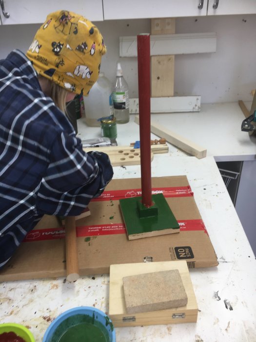 Barn i verkstad målar träprojekt med äggoljetempera: en leksakstelefon och en toapappersrulle.