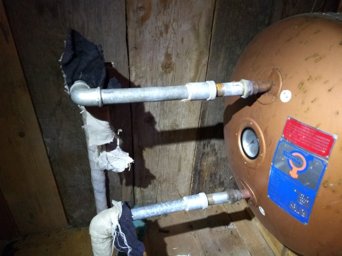 Varmvattenrör med läckage på skarv nära vattenberedare på en vind, delvis inslaget i trasig isolering.