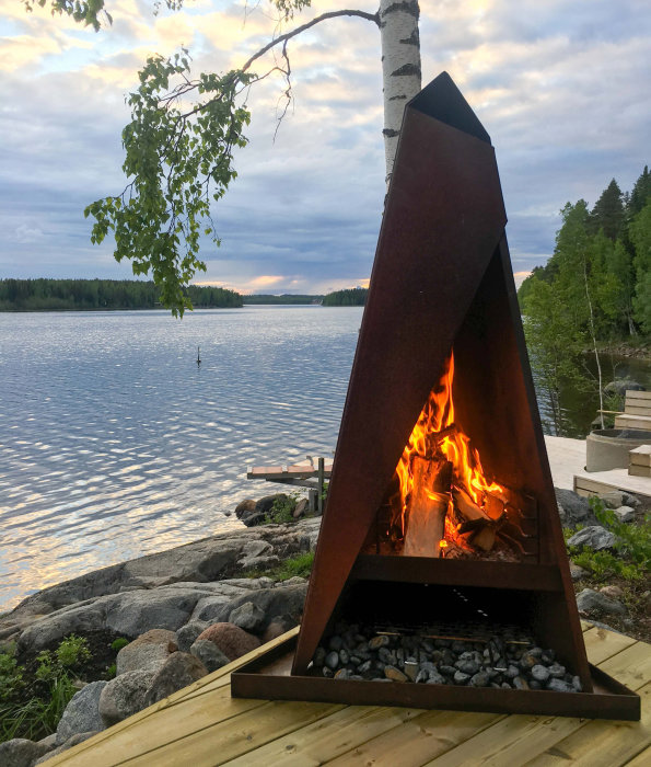 Vedeldad terrassvärmare i cortenstål vid sjö med brinnande eld vid skymning.