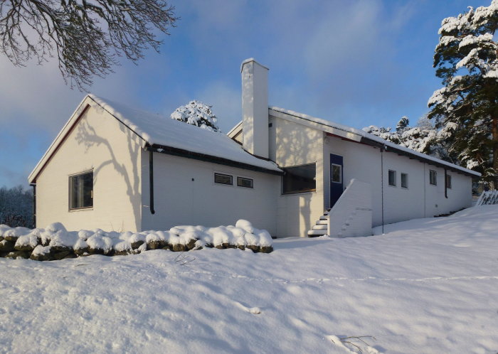 Vita sommarhuset Stennäs i två delar under vintern med snötäckt tak och mark, designat av arkitekten Gunnar Asplund.