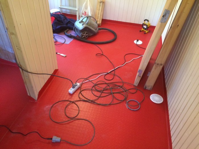 Röda plastmattor på golv under installation med verktyg, kablar och dammsugare i ett rum under renovering.