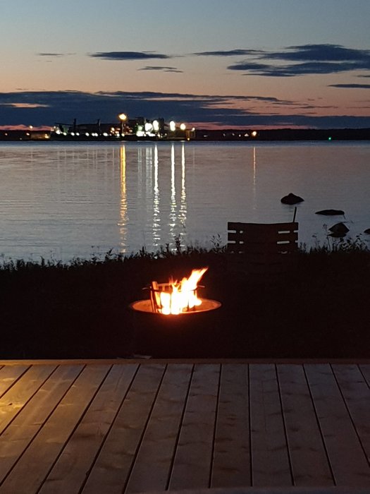 Eld brinner i en utomhuseldstad vid sjö med fabrik i bakgrunden under skymning.