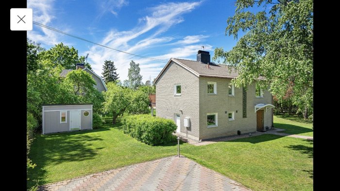 Tvåvåningshus med mexitegel-fasad omgiven av en välskött gräsmatta och lövträd under klarblå himmel.